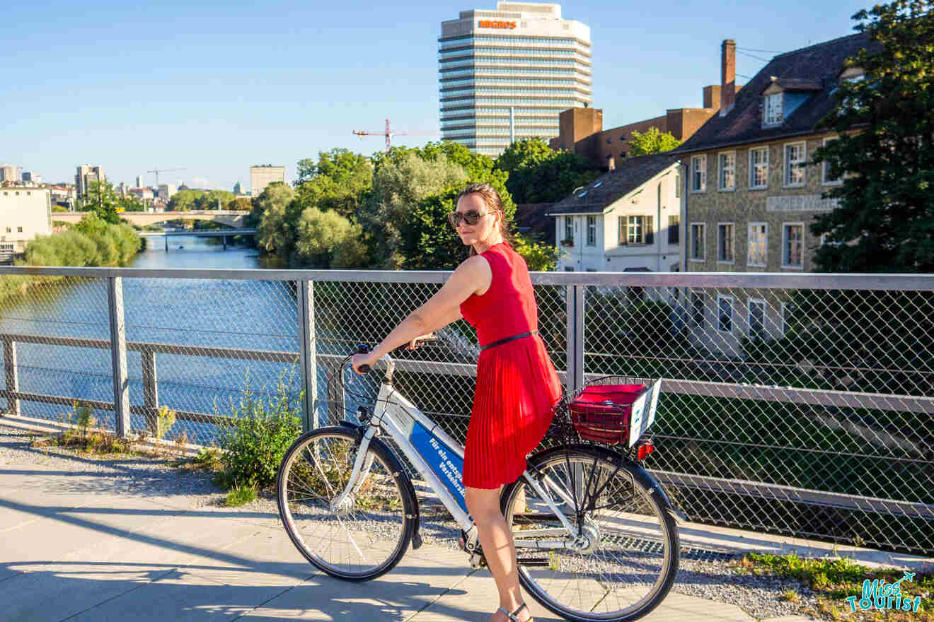 Yulia, the founder of this website, in a red dress stands with a bicycle on a riverfront path, with trees, buildings, and a high-rise visible in the background.