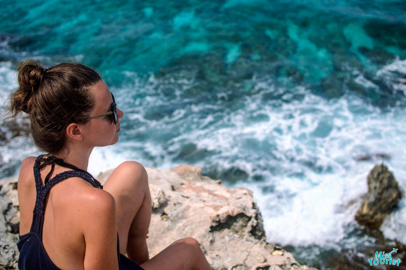 author of the post with sunglasses sits on a rocky cliff overlooking a turquoise ocean with waves crashing below.