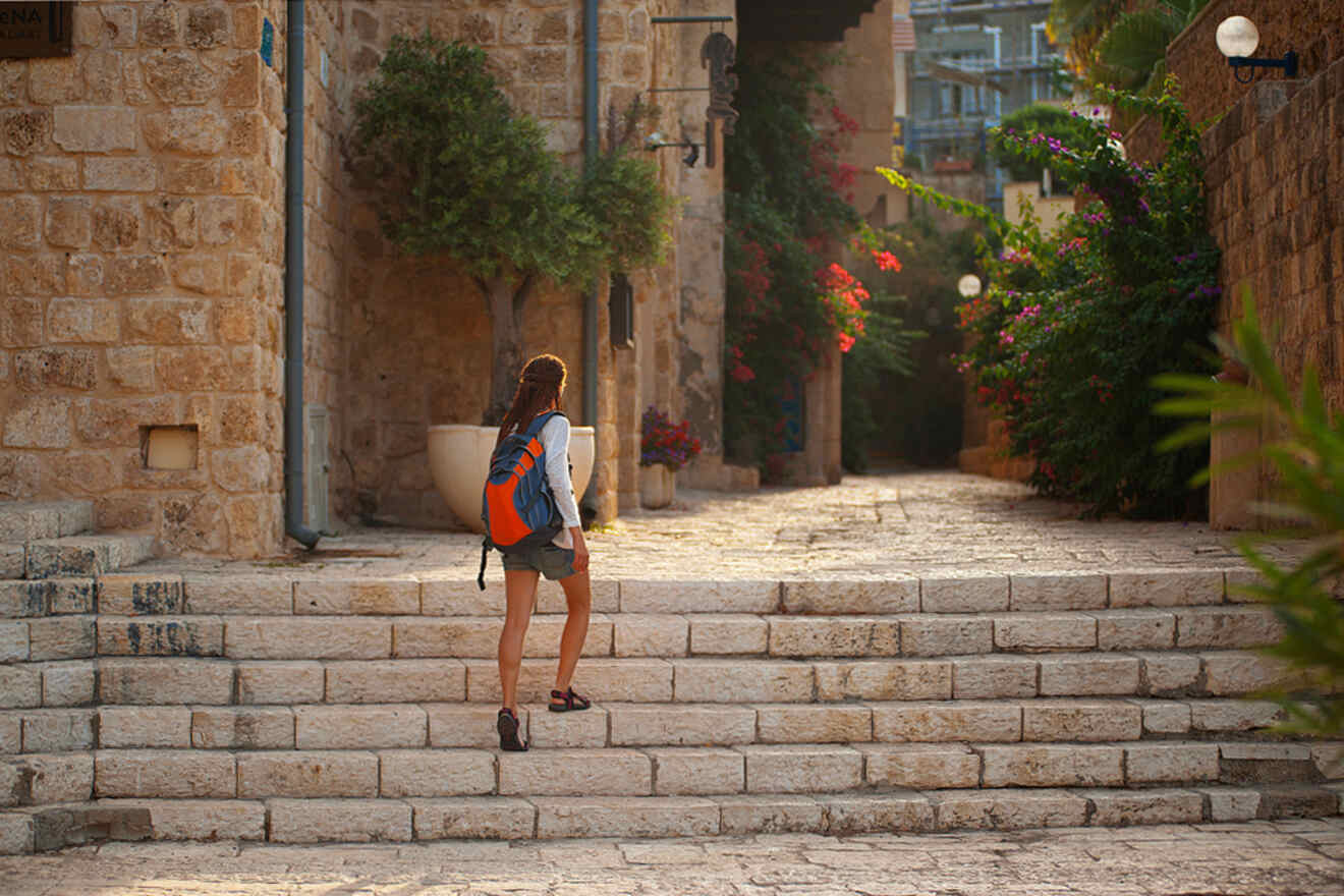 A person with a backpack walks up stone steps in a sunlit alley lined with plants and flowers.