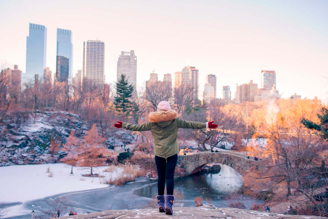 Person facing a cityscape with outstretched arms, standing in a snowy park with a stone bridge and trees in the background.