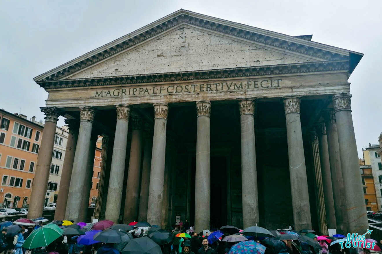 The Pantheon in Rome, with a crowd holding colorful umbrellas in front, on a cloudy day.