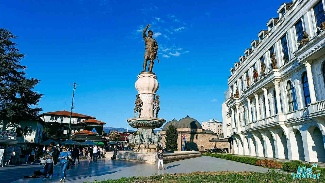 A large statue of a historical figure stands atop a fountain in a public square surrounded by buildings, under a clear blue sky.