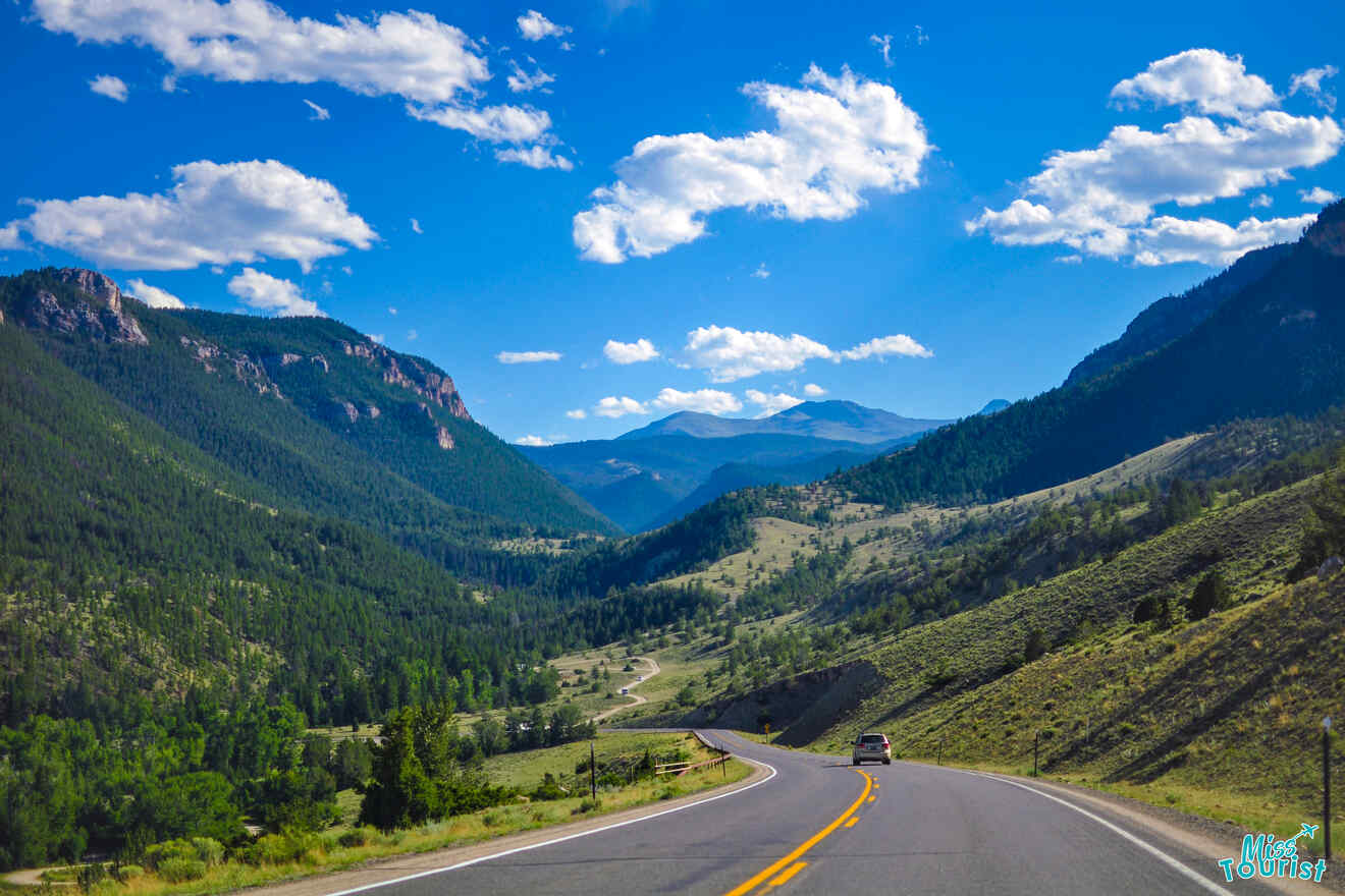 A winding road leads through a mountainous landscape with green hills and a bright blue sky with scattered clouds. A single car drives along the road.