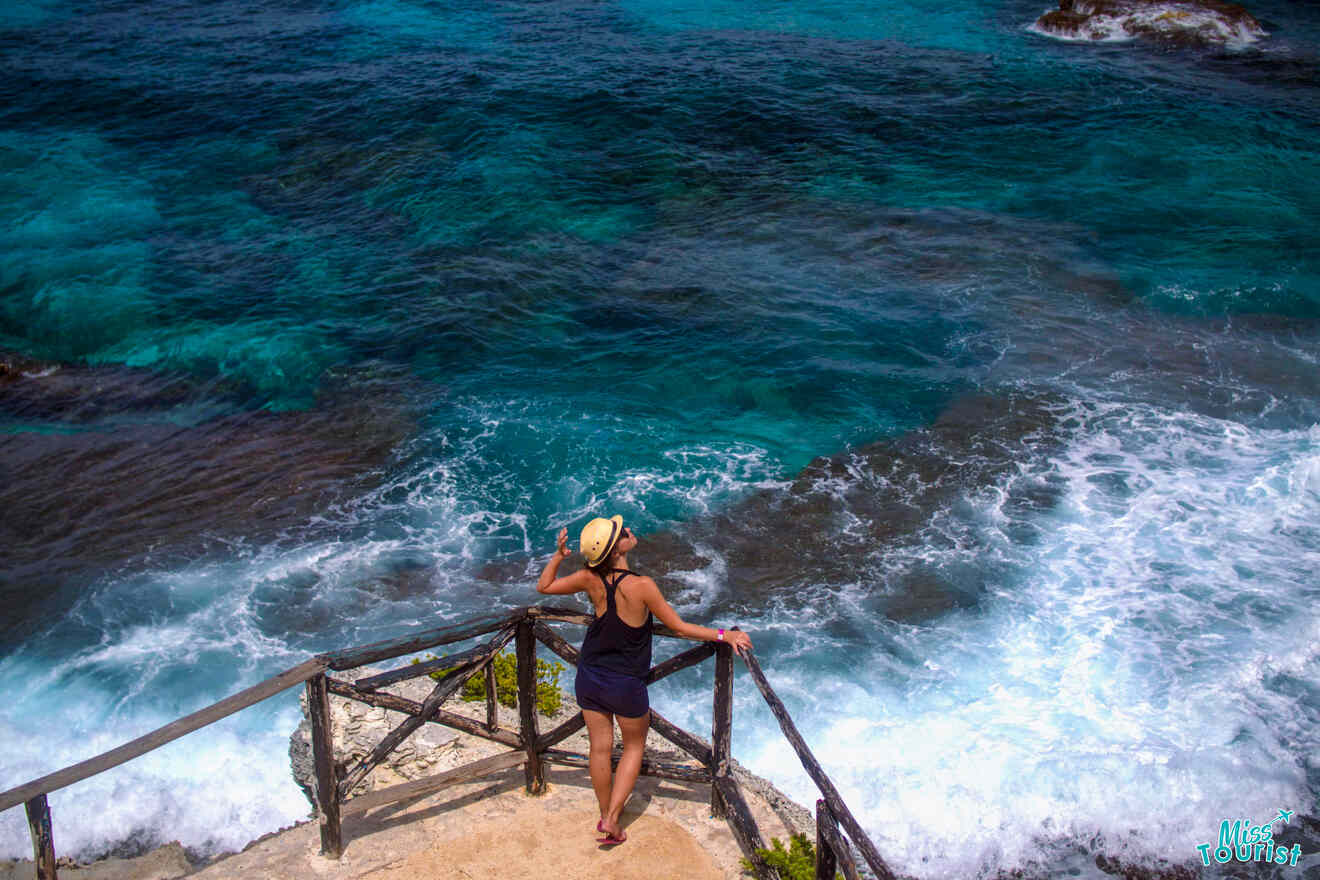 author of the post in a black swimsuit and hat stands on wooden stairs overlooking a turbulent blue ocean.