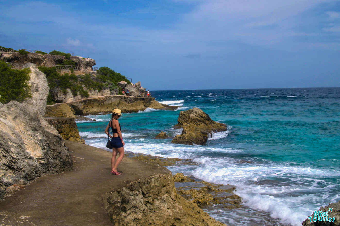 author of the post in a hat stands on a rocky coastal path, overlooking a bright blue sea under a clear sky.