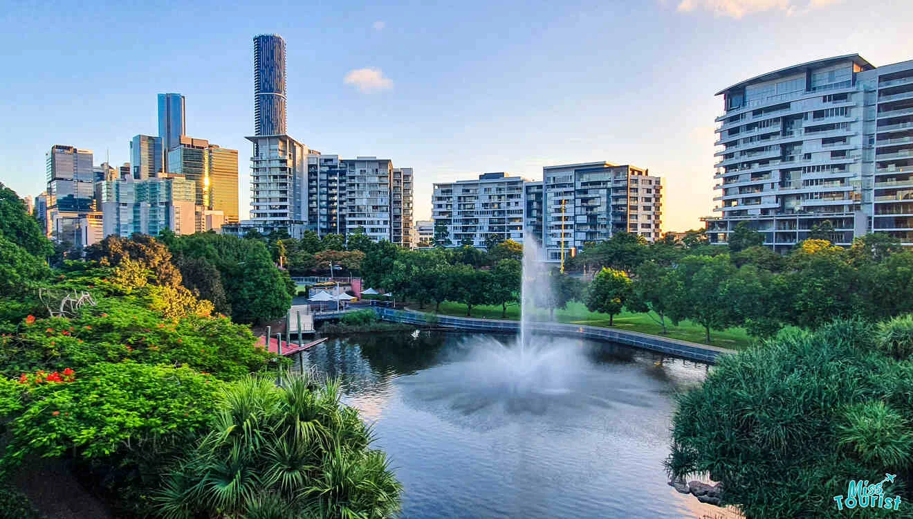 Cityscape featuring modern high-rise buildings and a central fountain in a park with greenery and a pond.