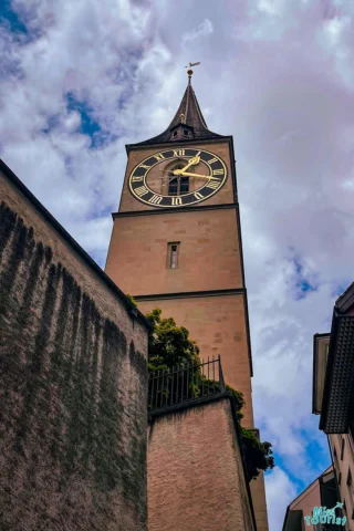 A tall clock tower with a sharp spire against a cloudy sky, surrounded by buildings and green foliage.