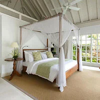 A bedroom with a four-poster bed draped in white fabric, green accent pillows, a side table with a lamp and flowers, and large windows letting in natural light.