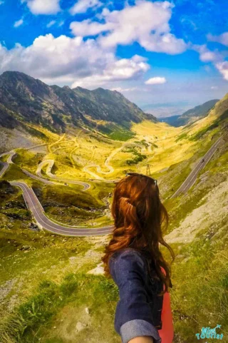 Yulia, the founder of this website, with long hair taking a selfie on a mountain road with winding paths, surrounded by grassy hills under a partly cloudy sky.