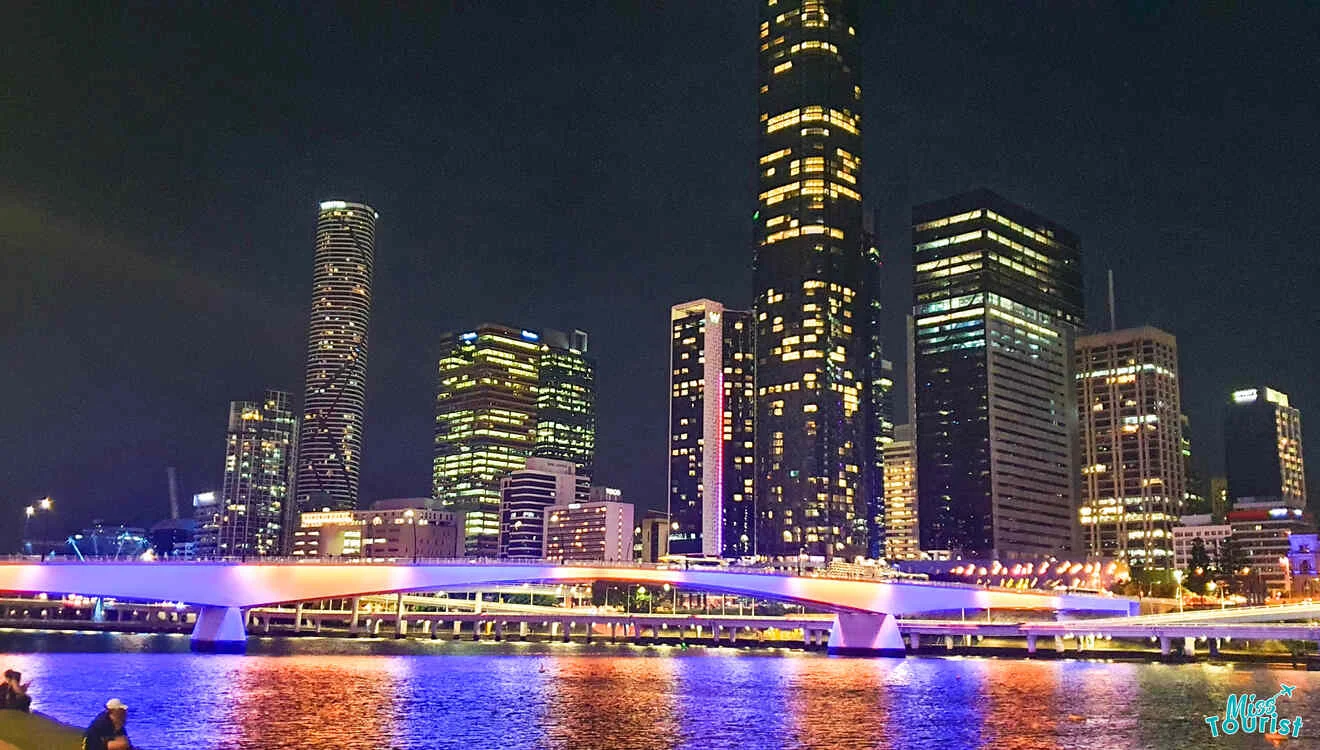 A brightly lit city skyline at night with tall skyscrapers and a bridge reflecting on the water.