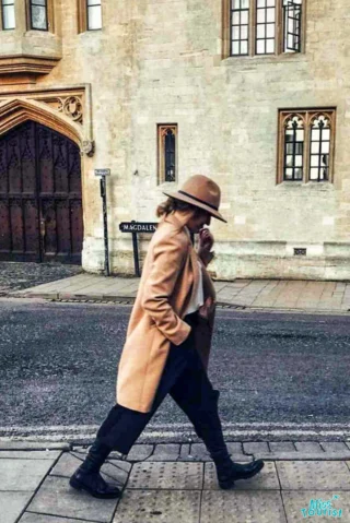 author of the post in a tan coat and hat walks along a sidewalk next to a stone building with arched windows and a wooden door, with a street sign in the background.