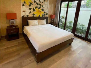 Uma cama impecavelmente arrumada com lençóis brancos ocupa o centro de um quarto com piso de madeira, janelas amplas e um mural floral na parede atrás da cabeceira.