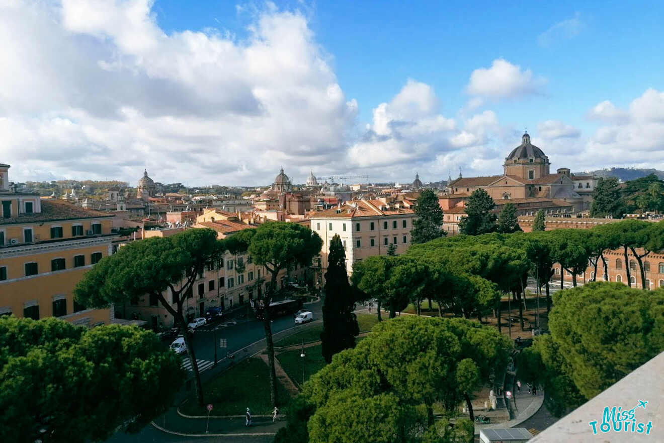 View of a cityscape with historical buildings, lush green trees, and a partly cloudy sky.