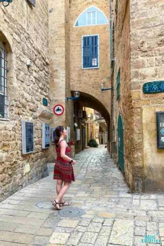 The writer of the post in a red dress stands in a narrow, stone-paved alleyway lined with historic stone buildings.