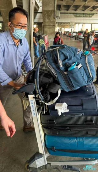 A person wearing a mask pushes a luggage cart loaded with multiple suitcases and bags at an airport terminal.