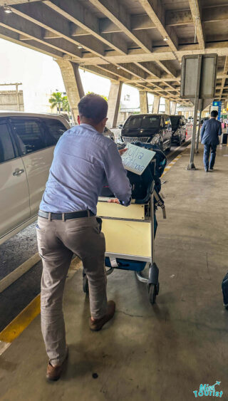 Person pushing a luggage cart under a concrete structure, with a sign on the cart. Cars are parked nearby.