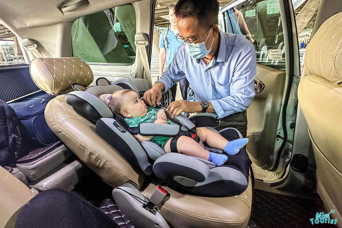 A man secures a baby in a car seat inside a vehicle. The man is wearing a mask, and the baby is looking up at him.