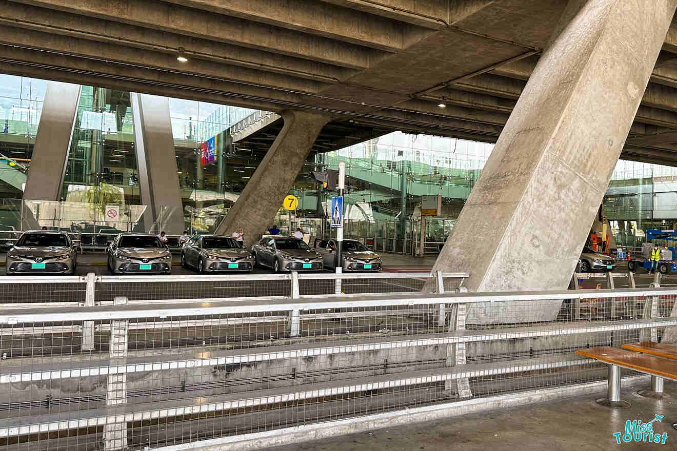 Line of parked taxis under a large concrete structure at an airport terminal, with glass buildings in the background.