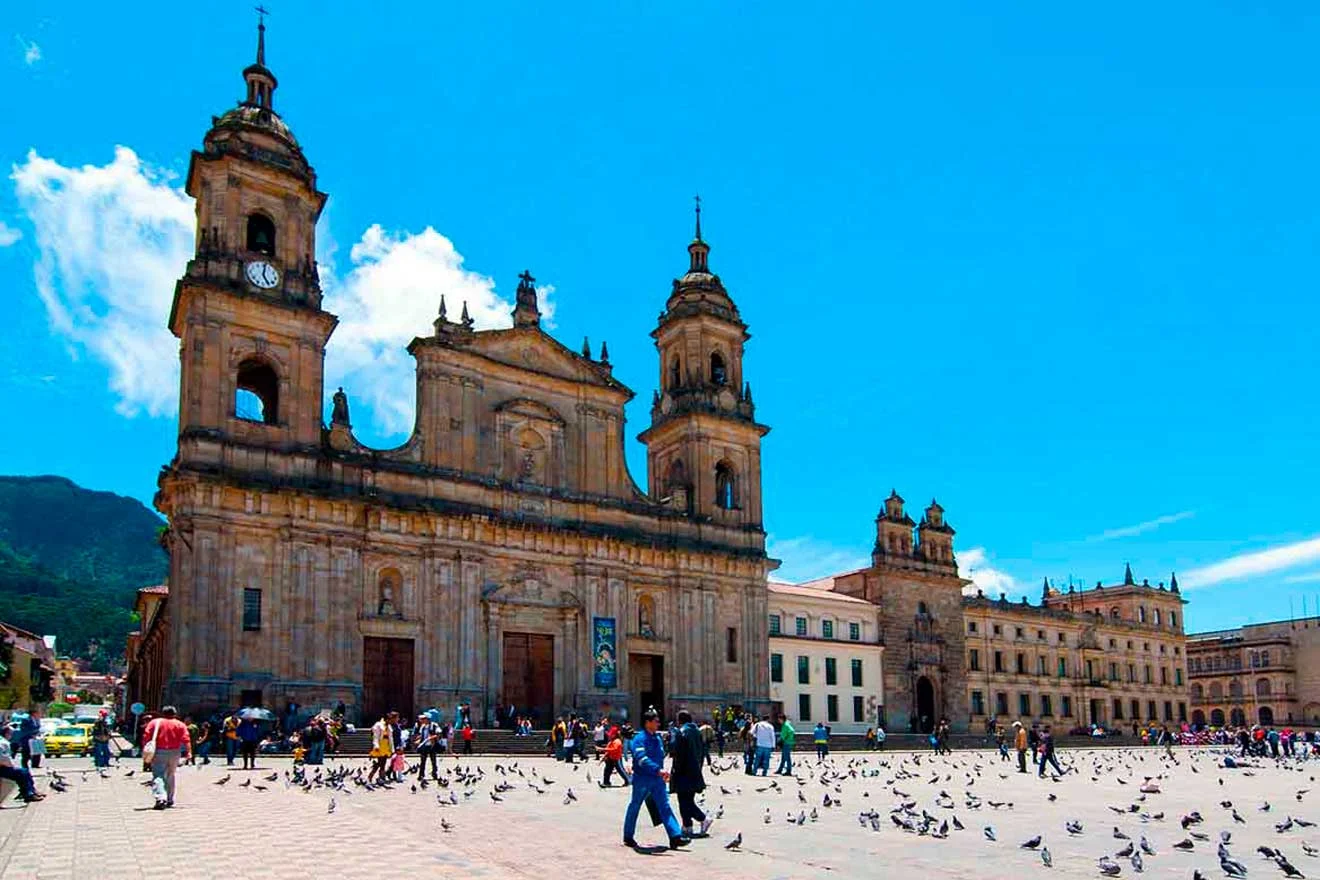 A bustling city square with a large historic cathedral, numerous people, and pigeons on a sunny day.