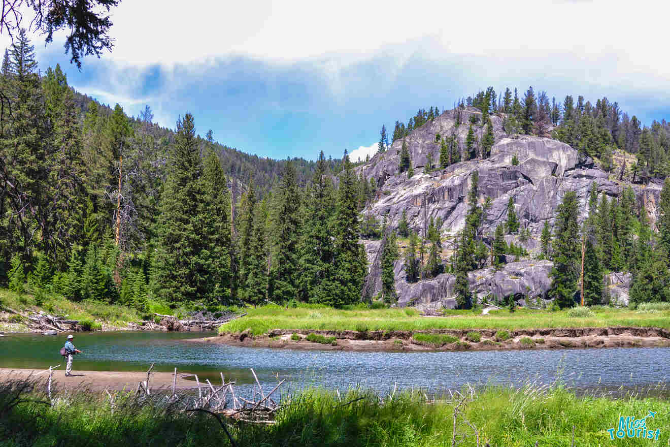 A person stands by a serene river, surrounded by lush green trees and rocky hills under a partly cloudy sky.