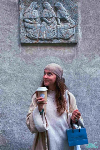 author of the post stands holding a coffee cup and a small blue shopping bag, looking upwards with a smile. Behind them is a stone relief of three seated figures.