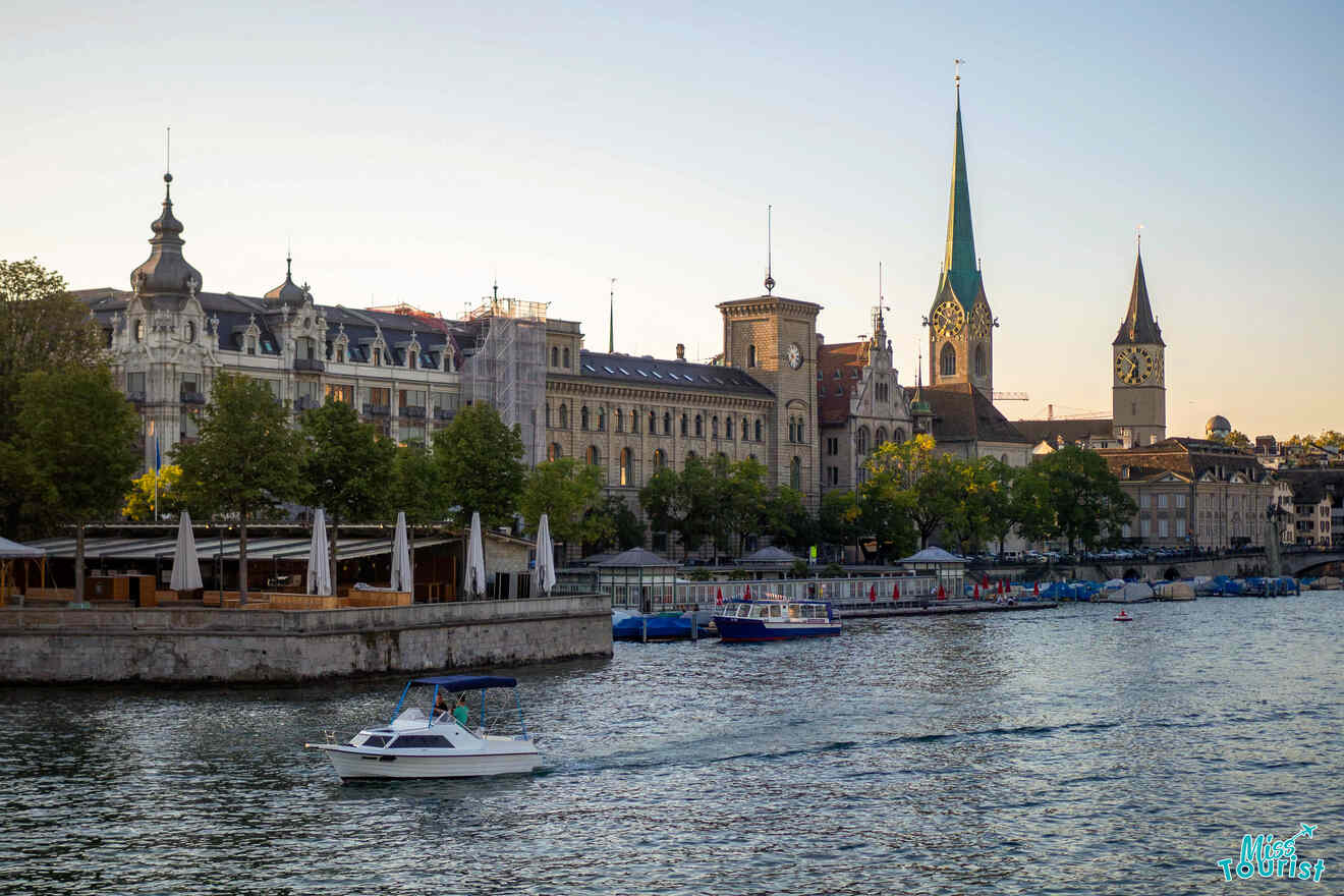 A small boat sails on a river with historic buildings and church spires in the background under clear skies.