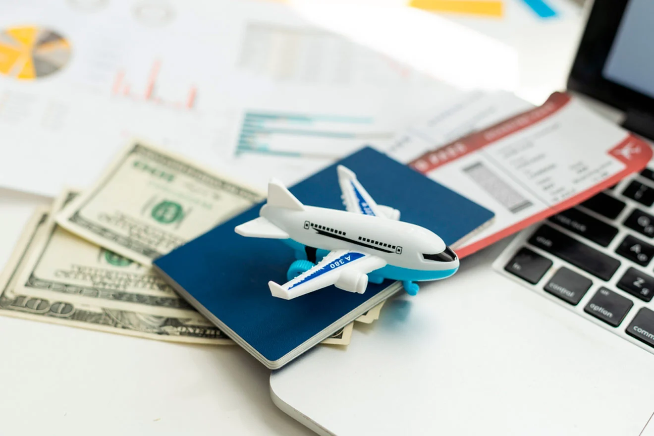 A toy airplane on a passport, beside cash, a boarding pass, and a laptop, all placed on a table with charts.