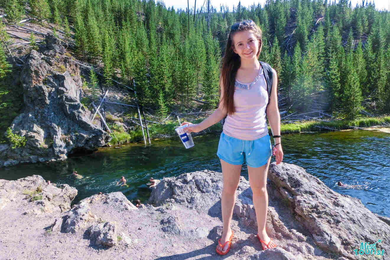 author of the post standing by a river in a forested area, holding a bottle. Swimmers are in the water behind her.