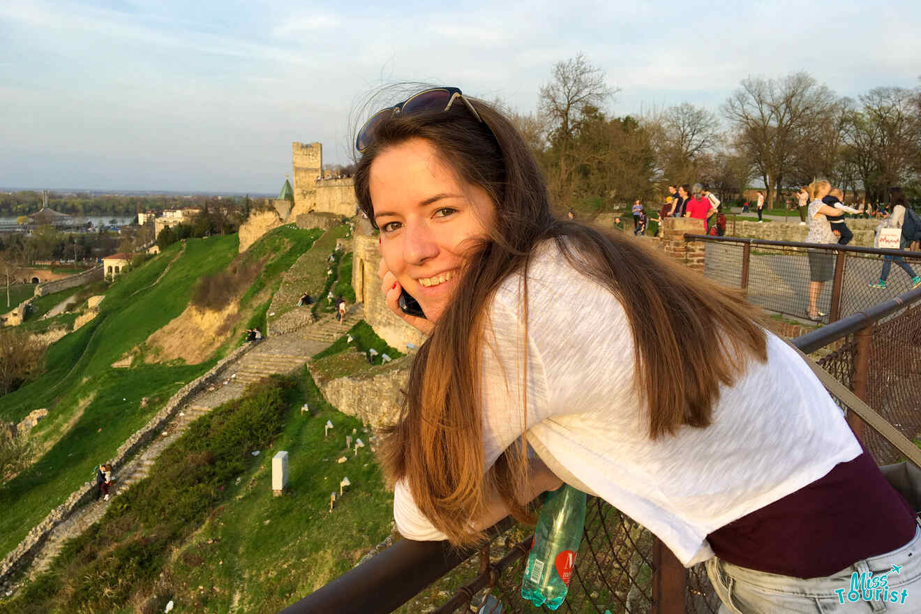 Woman smiling and leaning on a railing at a scenic viewpoint with a historic building and greenery in the background.