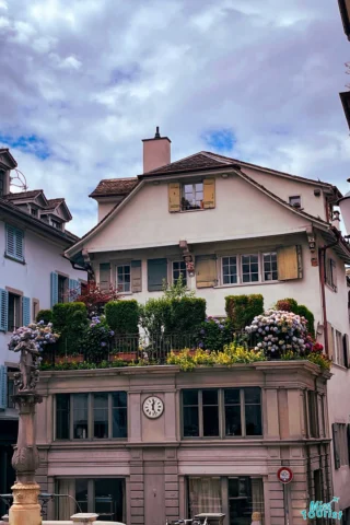 A historic building with a clock and lush rooftop garden, set against a cloudy sky.