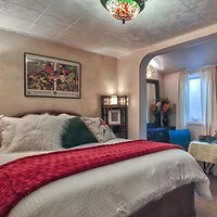 Cozy bedroom with a white bed, red throw blanket, decorative pillows, and a stained glass ceiling light. Features a small seating area with a window and colorful artwork on the wall.