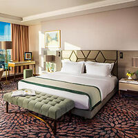 A modern hotel room with a large bed, upholstered bench, desk, and window with a city view. Neutral colors and patterned carpet adorn the space.