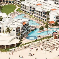 Aerial view of a beach resort with several pools, white buildings, palm trees, and beach chairs arranged along the sandy shore.