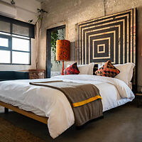 A modern bedroom with a double bed, colorful pillows, a large geometric wall art above the bed, a window on the left, and an orange lamp.