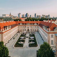 Aerial view of a historic building with red roofs and a formal garden courtyard, surrounded by trees. In the background, a city skyline with modern buildings is visible.