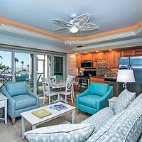 Modern living room with blue and grey seating, a dining set, and an open kitchen. A ceiling fan is above, and glass doors lead to a balcony with a view of palm trees and the ocean.