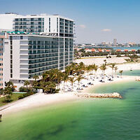 A modern beachfront hotel with balconies overlooks a sandy beach and green-blue water. Palm trees and lounge chairs line the shore under a clear blue sky.