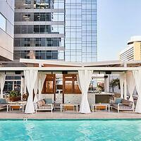 Rooftop pool area with white cabanas, modern loungers, and a bar, surrounded by tall city buildings.