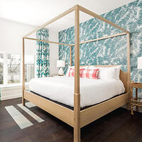 A bedroom with a wooden canopy bed, white bedding, and red plaid pillows. The room features a blue patterned accent wall and large windows on the left, with a small wooden side table.