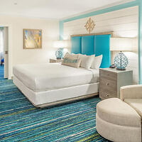 Bright hotel room with a large bed, blue accent wall, and colorful carpet. Features include two bedside tables with lamps and a decorative wall art.