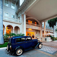 A vintage blue car parked in front of an elegant hotel entrance with arches and balconies.