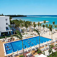 Aerial view of a beachfront resort with a large outdoor pool, surrounded by lounge chairs and palm trees, adjacent to the ocean.