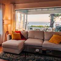 Cozy living room with a beige sectional sofa, yellow and red cushions, a floor lamp, and a large window showing a seaside view.