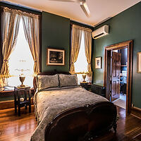 A bedroom with green walls features a double bed, draped windows, a ceiling fan, wall art, and a wooden floor. A door leads to another room.