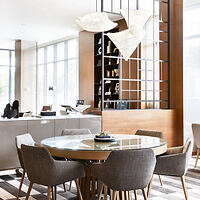 Modern dining area with a round glass table, six gray upholstered chairs, a decorative light fixture, and a wood and glass partition in the background.