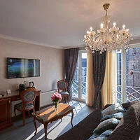 A living room with a chandelier, wall-mounted TV, sofa, wooden table, two chairs, and a bouquet of flowers. Large windows with curtains allow light to fill the room.