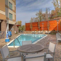 Outdoor pool area with a round table and chairs in the foreground, surrounded by a wooden fence and trees in the background.