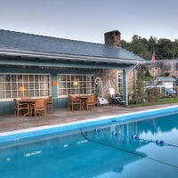 Outdoor swimming pool with blue water in front of a one-story building with large windows and wooden tables. An American flag is in the background.