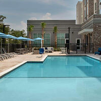 Outdoor swimming pool with clear water, surrounded by lounge chairs and blue umbrellas. Gray building with large windows and a brick section is in the background.