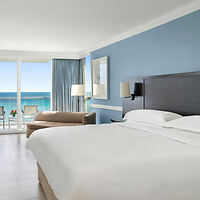 Hotel room with a large bed, ocean view through glass doors, blue walls, a sofa, and a lamp.